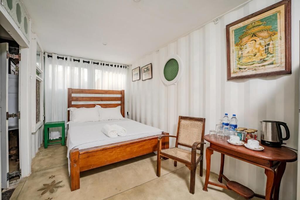 A neatly arranged bedroom with a wooden bed, a chair, a small table with a kettle, and a painting on the wall.