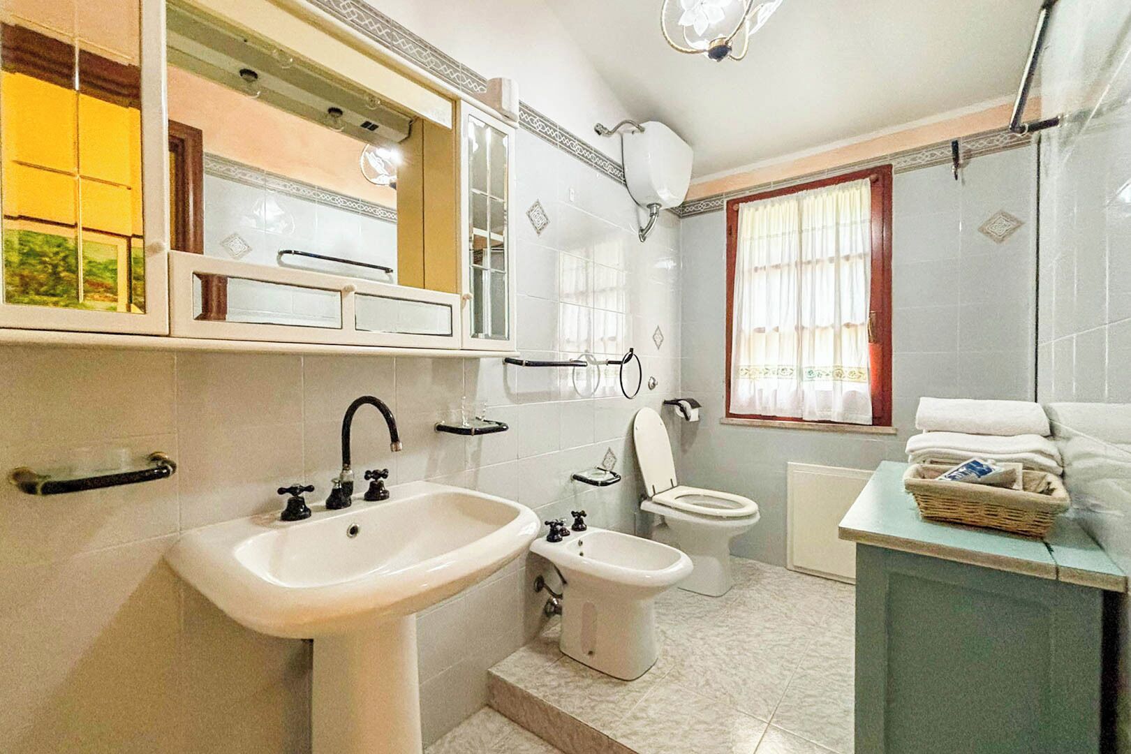 A bathroom with a sink, bidet, toilet, and a window with curtains.