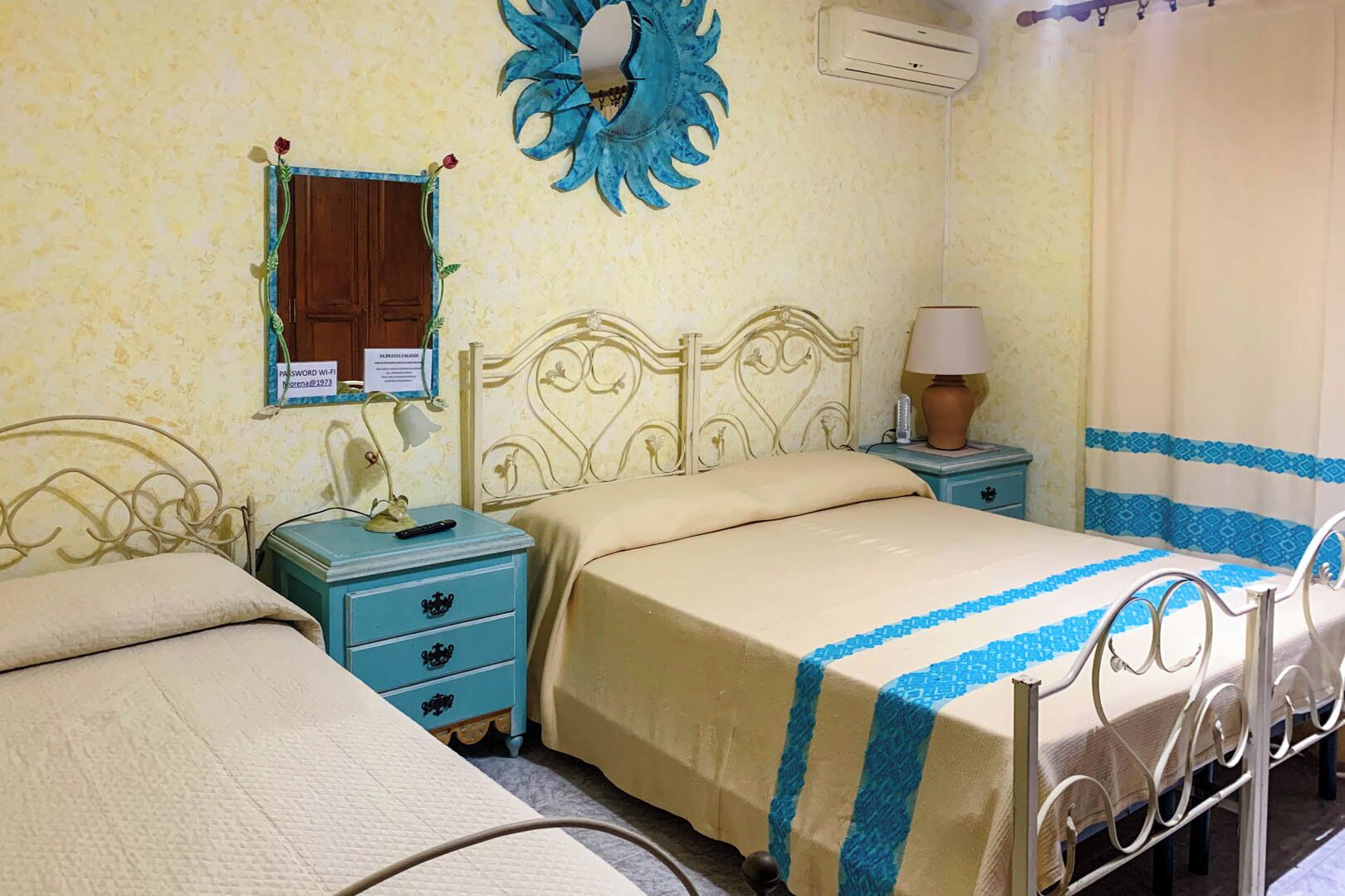 A bedroom with two beds, a nightstand with a lamp, a wall-mounted air conditioner, and a decorative wall piece.