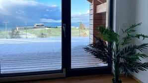 A room with a large sliding glass door leading to a wooden deck, a potted plant, and a view of a building and landscape. - Eichen-Chalet (Eichenberg)