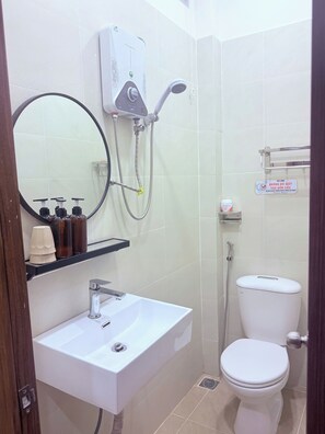 A small bathroom with a white sink, a toilet, and a shower. There is a mirror above the sink, and a soap dispenser is visible. A sign is mounted on the wall, and a small shelf is attached to the wall. - Sonder Hau Giang Hotel (Tan Binh)