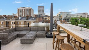 Una terraza en la azotea con mobiliario de mimbre, una mesa de madera y vistas a edificios de la ciudad.