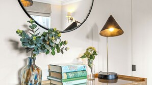 Un escritorio de madera con un espejo redondo, un florero con follaje verde, una lámpara y una pila de libros.