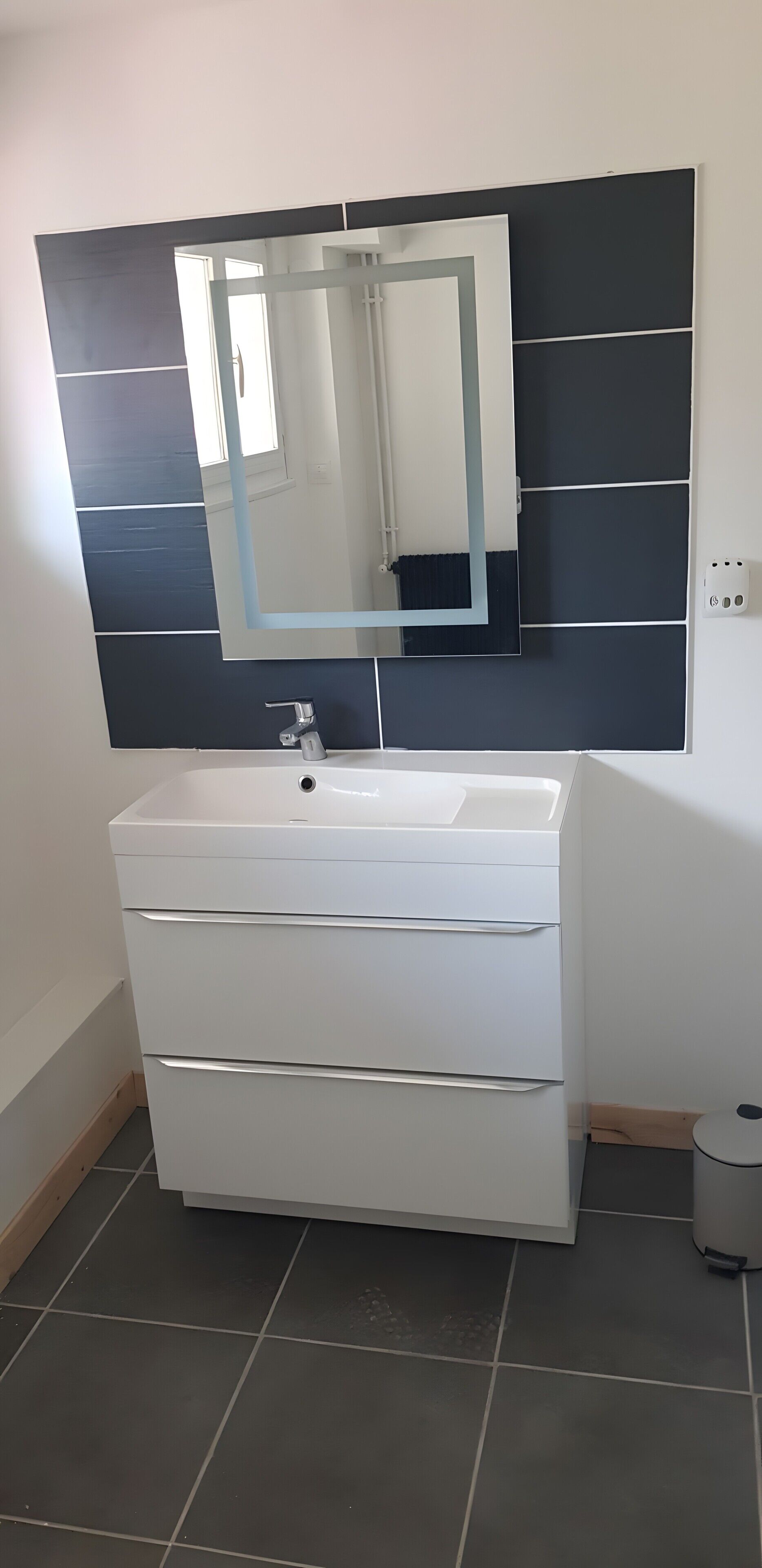 A bathroom with a white vanity, a large mirror, and a dark tiled backsplash.