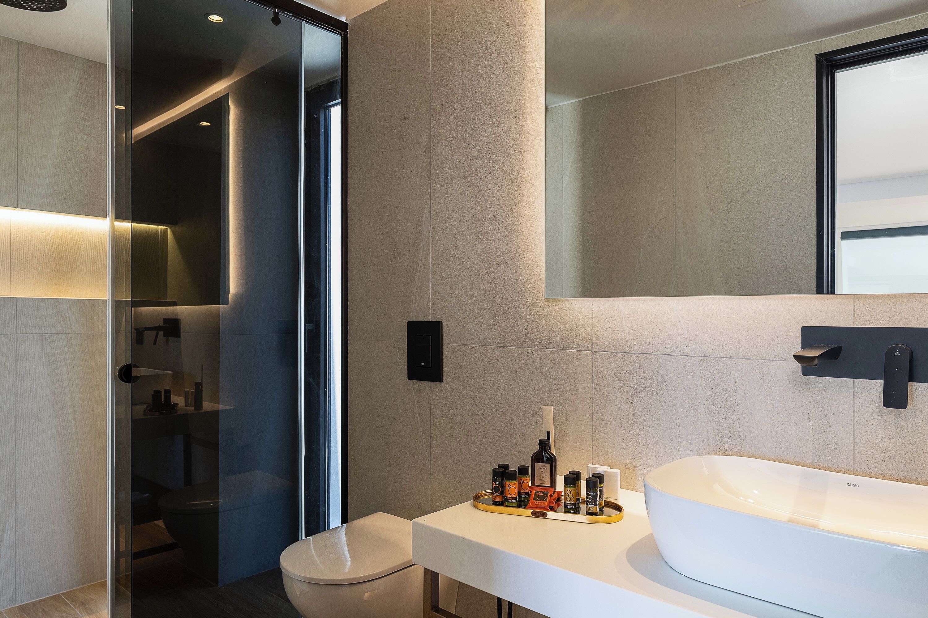 Un baño moderno con cabina de ducha de vidrio, un lavabo blanco y un atril con artículos de aseo, y un espejo grande.