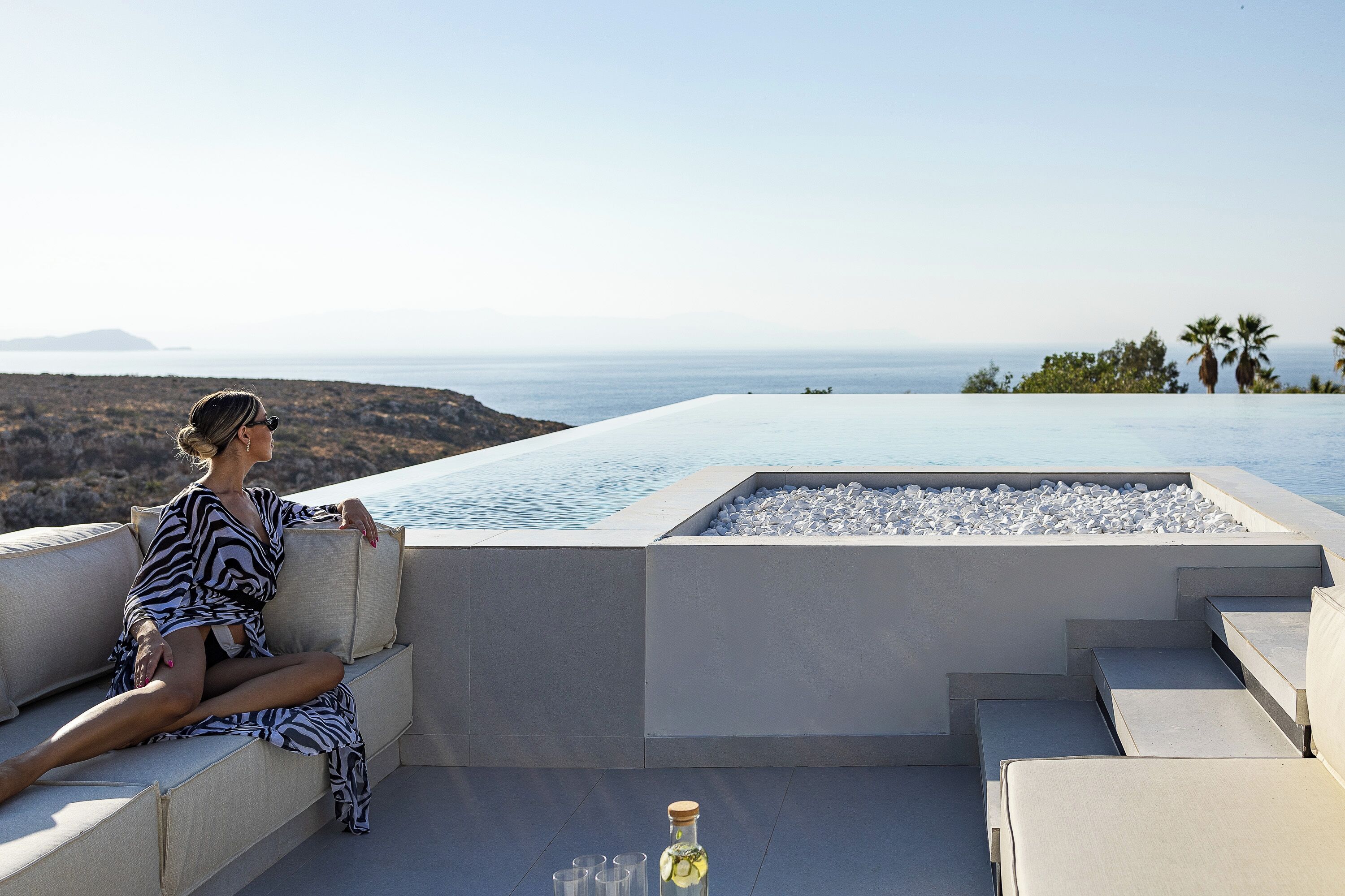 A woman sitting on a cushioned bench by a pool with a clear view of the ocean and distant hills.