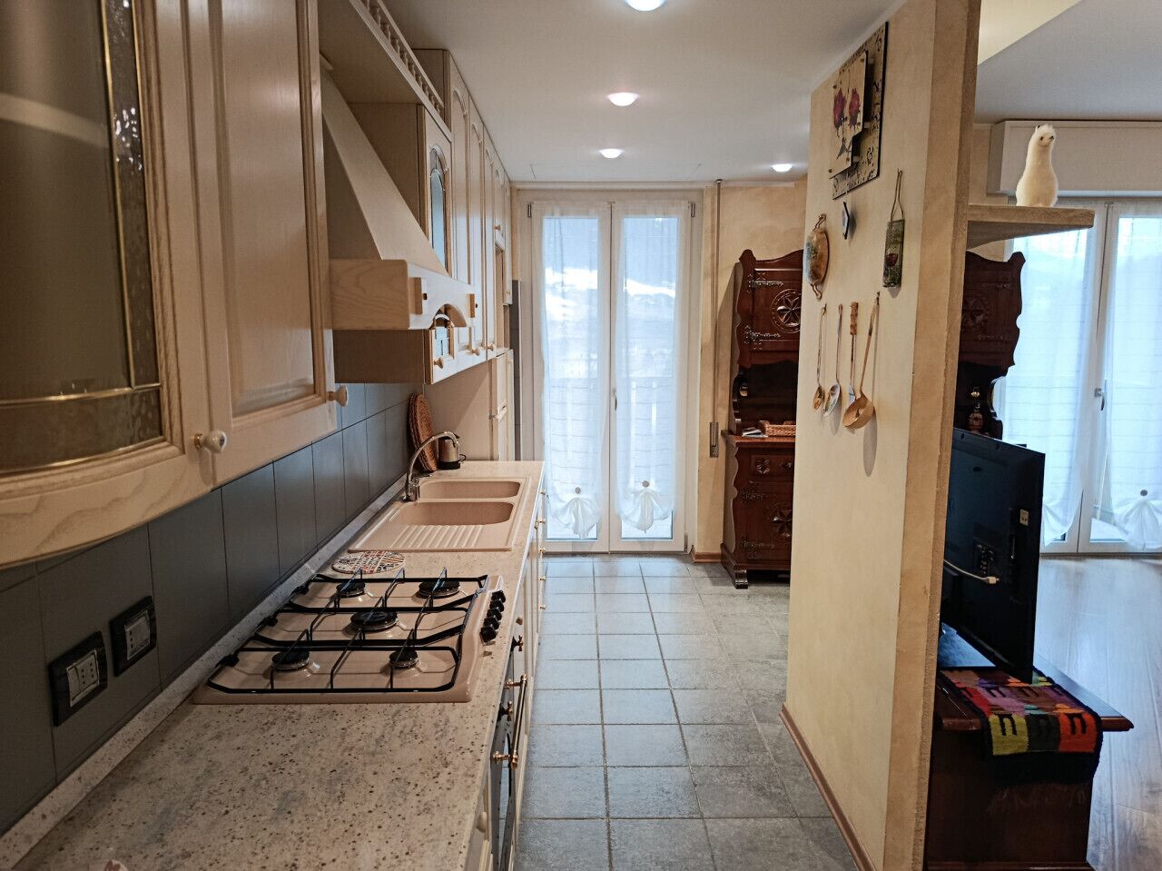 A kitchen with a gas stove, sink, and cabinets. There is a wooden cabinet with decorative items and a small table with a colorful cloth.