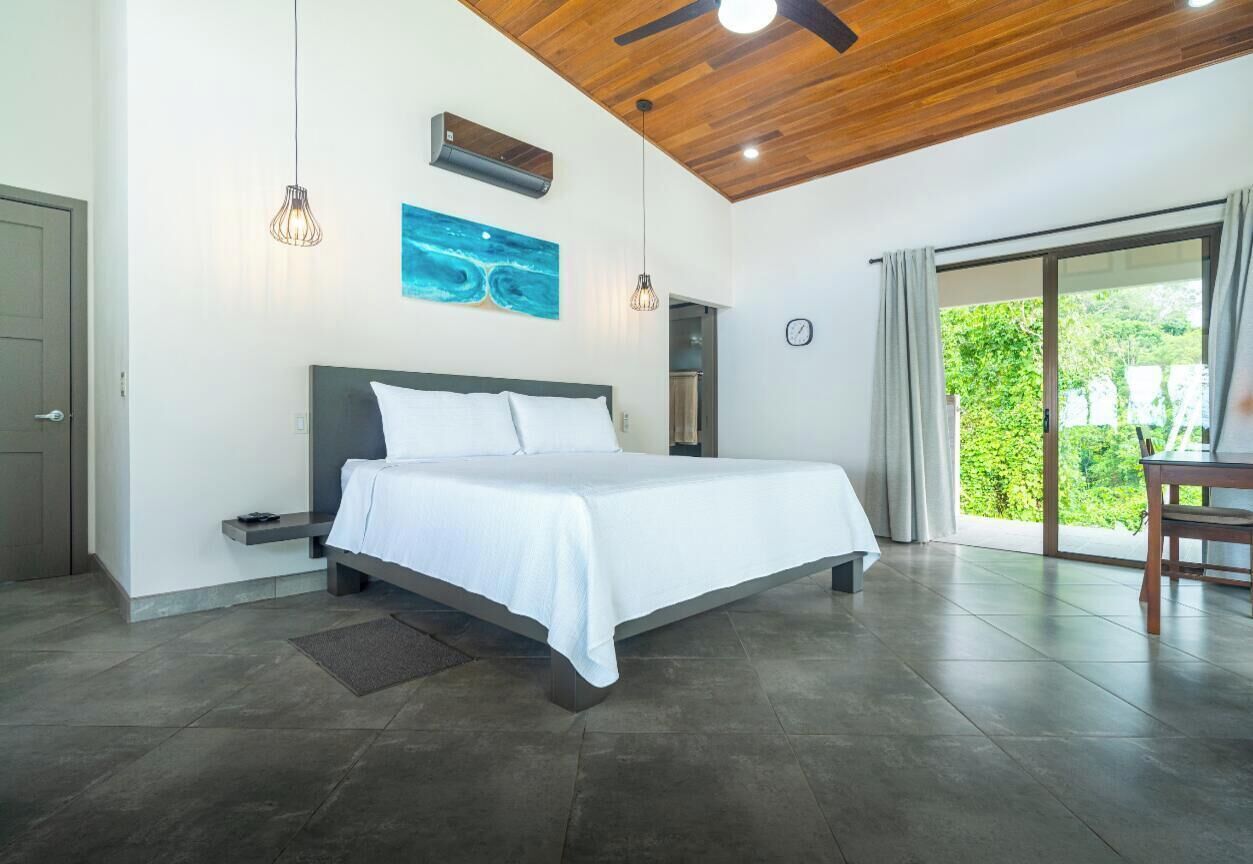 A modern bedroom with a large bed, a ceiling fan, and a view of greenery through the sliding glass door.