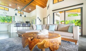 Un salon moderne avec une grande table basse en bois, un canapé beige agrémenté de coussins bleus, et une cuisine en arrière-plan.