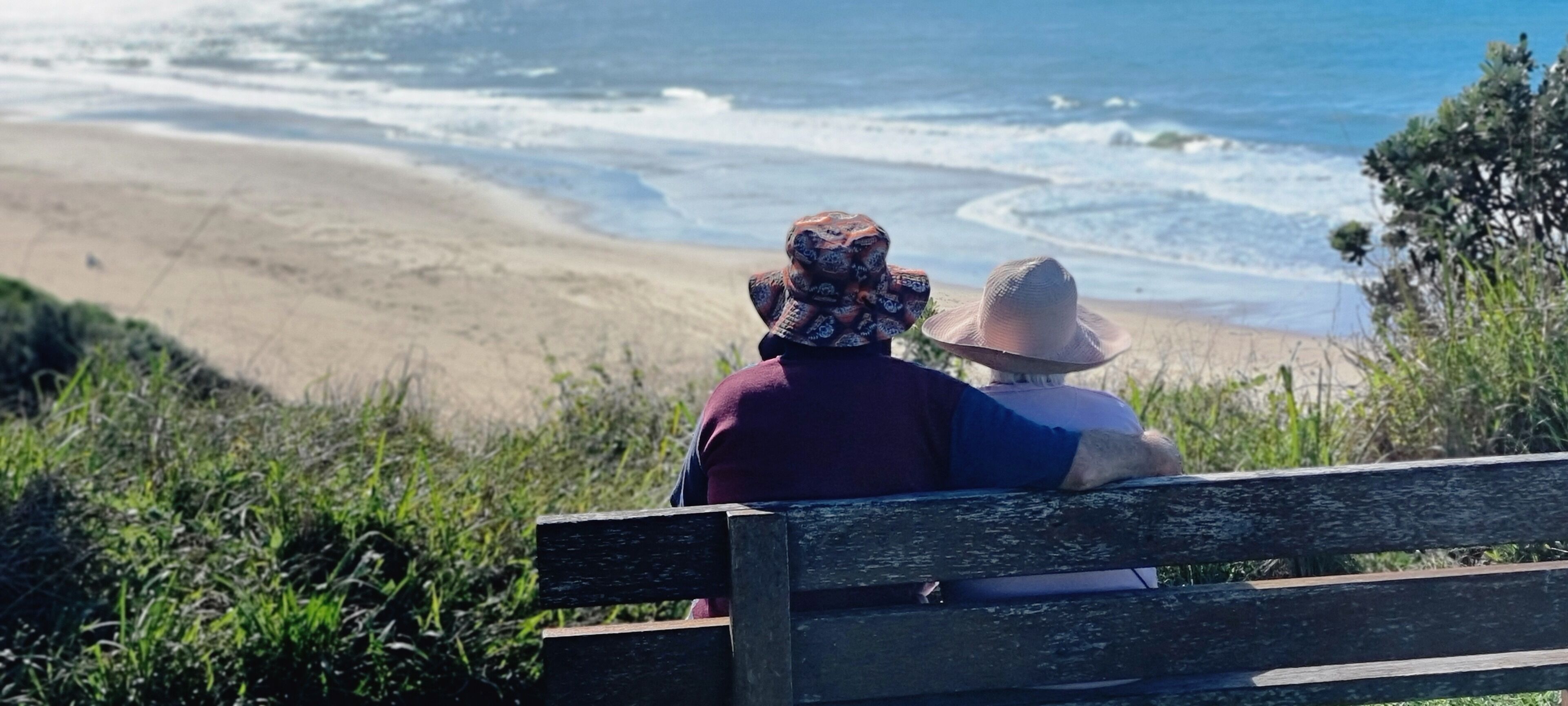 Two people sitting on a wooden bench overlooking a beach with waves.