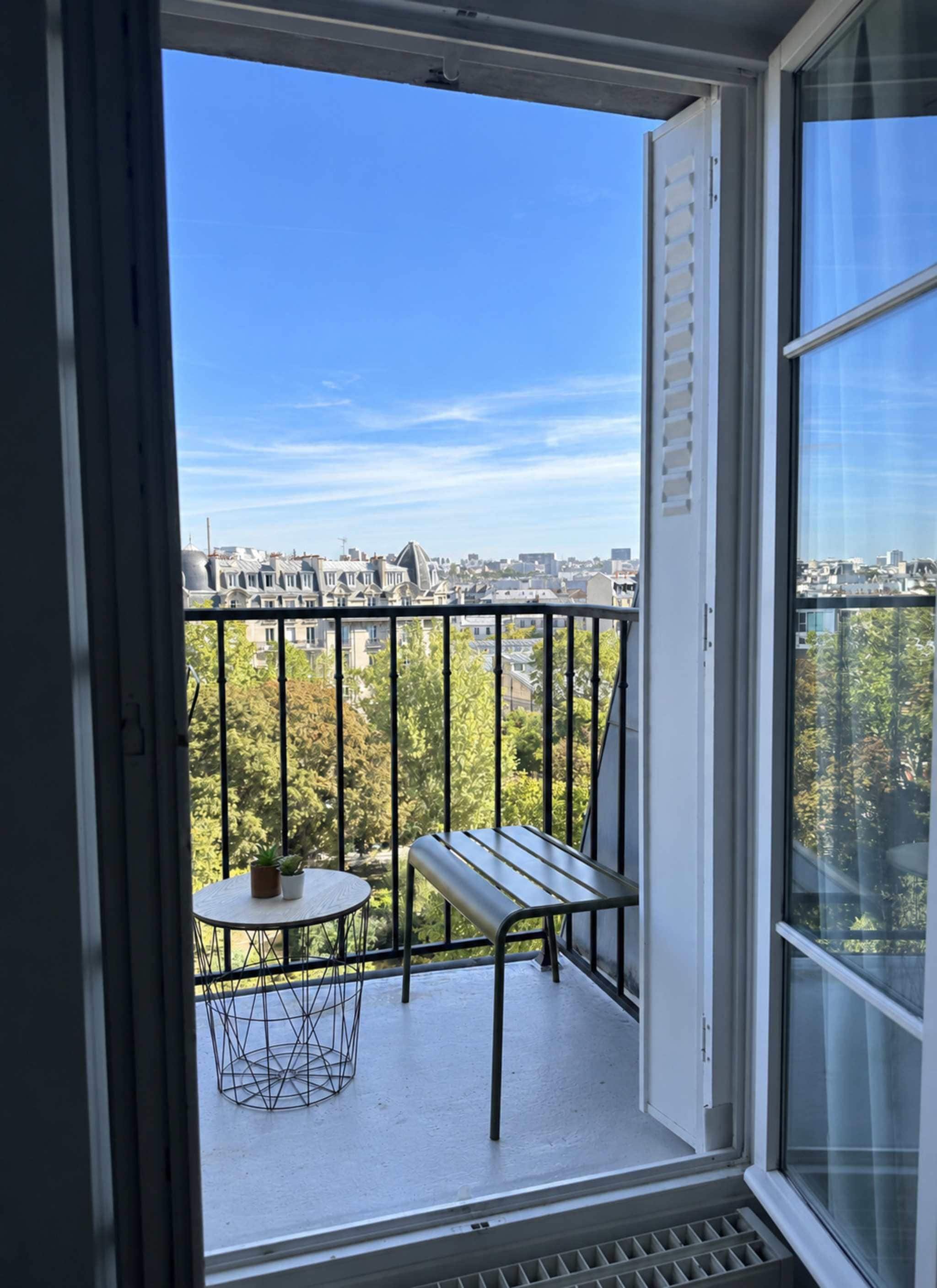 A balcony with a metal chair and a small table, a clear view of the cityscape, and a window with blinds.