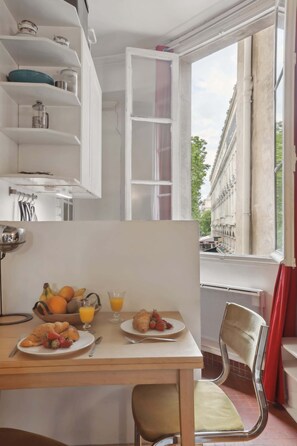 A breakfast table with croissants, fruit, and orange juice, a chair, and a view of a building through a window. - Chic Studio near La Seine & Rivoli (Paris)