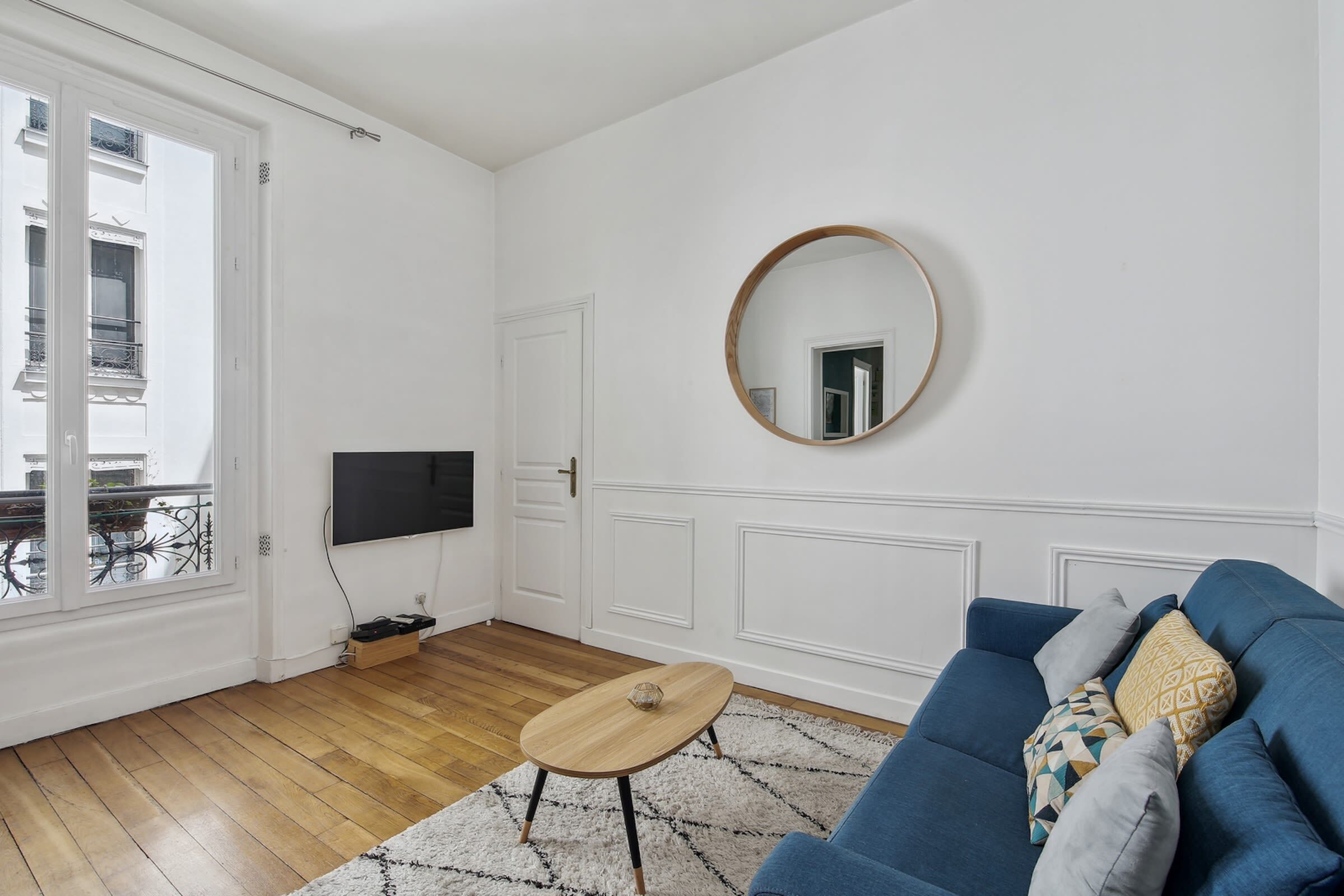 A living room with a blue sofa, a round mirror, a wooden coffee table, and a TV on the wall.