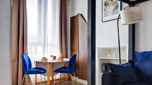 Ein Zimmer mit einem Holzespeisetisch, zwei blauen Samtstühlen, einem dunkelblauen Sofa, einem weißen Regal mit Dekoration und einem Gemälde an der Wand.