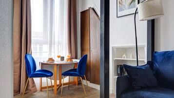 Una habitación con una mesa de comedor de madera, dos sillas de terciopelo azul, un sofá azul oscuro, un estante blanco con adornos y un cuadro en la pared.