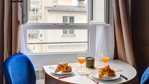 Ein Zimmer mit einem runden Frühstückstisch, zwei blauen Stühlen, einem Fenster mit Vorhängen und Blick auf Gebäude.