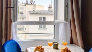 Una habitación con una mesa redonda dispuesta para el desayuno, dos sillas azules, un ventanal con cortinas y vistas a edificios.