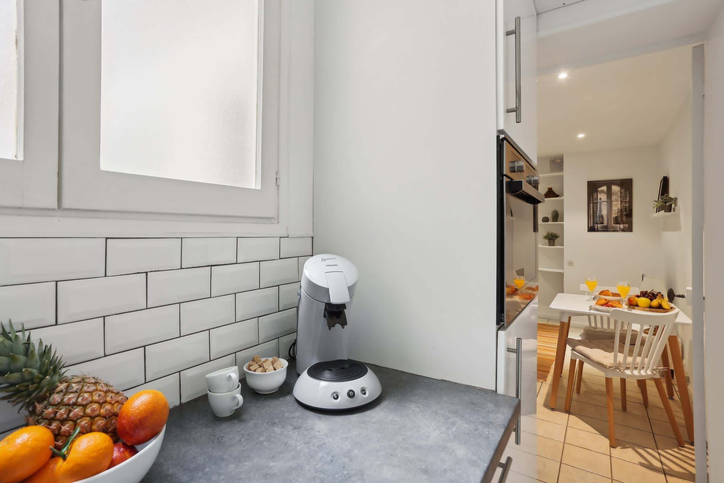 A kitchen with a coffee machine, a bowl of fruit, and a dining area with a table and chairs.