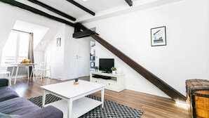 A modern living room with a white sofa, a coffee table, and a TV on a white cabinet. A wooden staircase leads to an upper level. - Le Marais - 1BR by Pompidou (Paris)