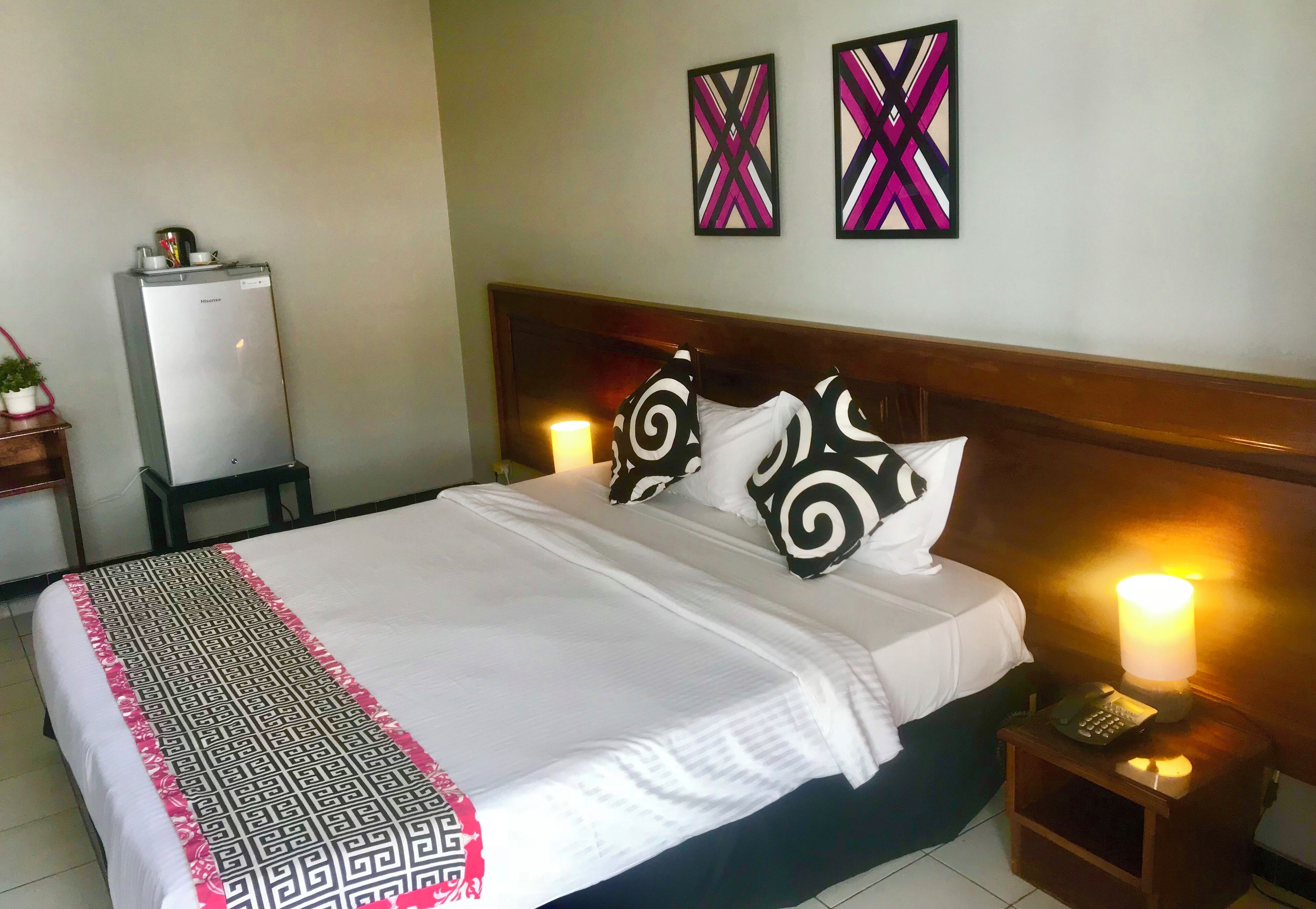 A neatly made bed with a patterned bedspread, two wall-mounted abstract paintings, a bedside lamp, and a small table with a phone.