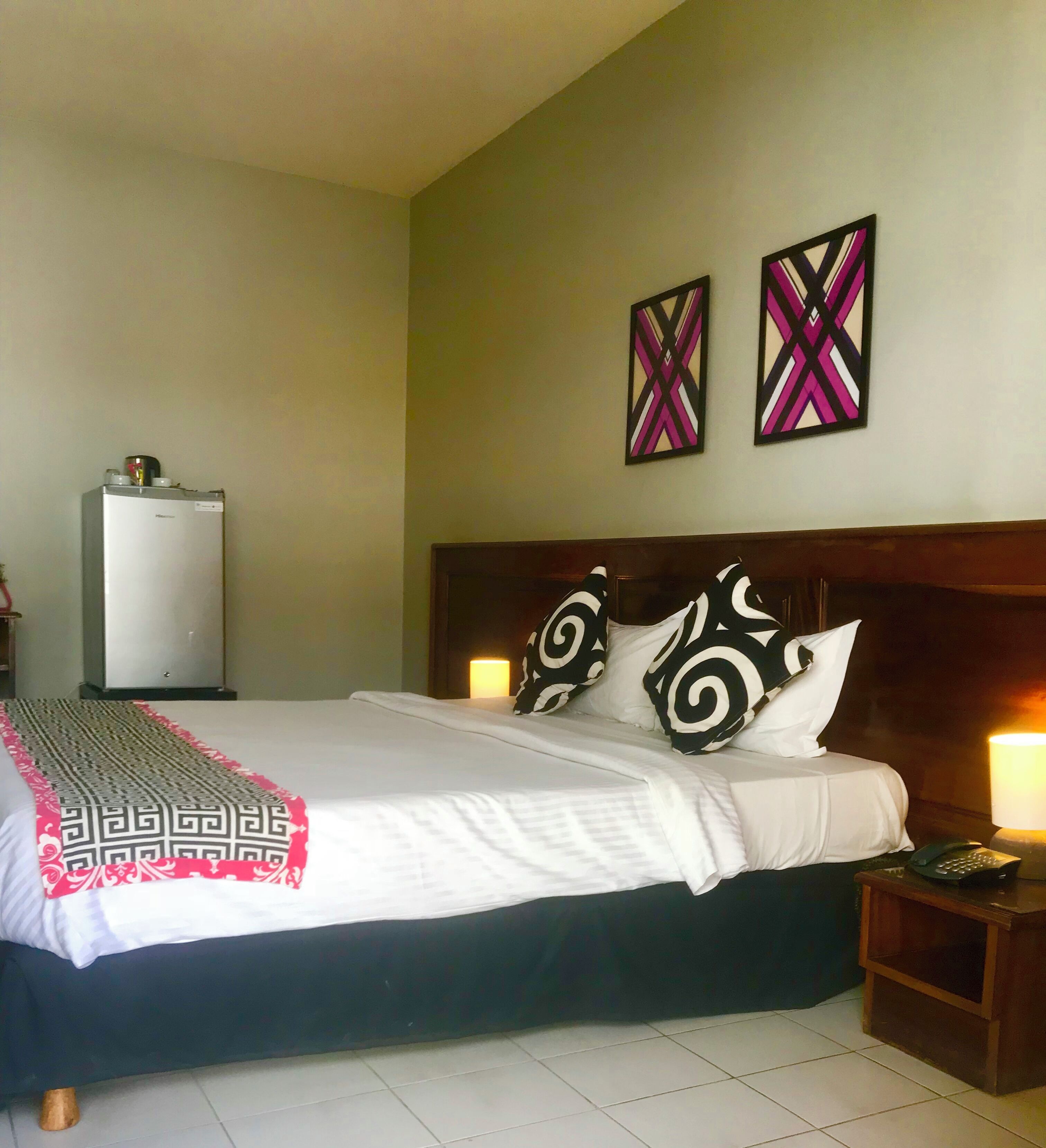 A hotel room with a bed, bedside lamps, a nightstand, a refrigerator, and two wall art pieces.