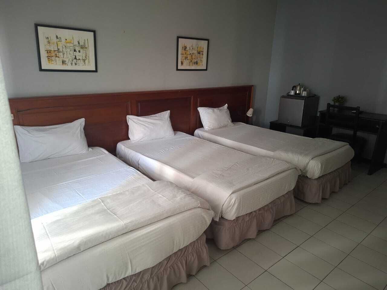 Ein Hotelzimmer mit drei Einzelbetten, einem Holzkopfteil, zwei gerahmten Bildern an der Wand, einem kleinen Tisch mit einem Wasserkocher und einem Stuhl.