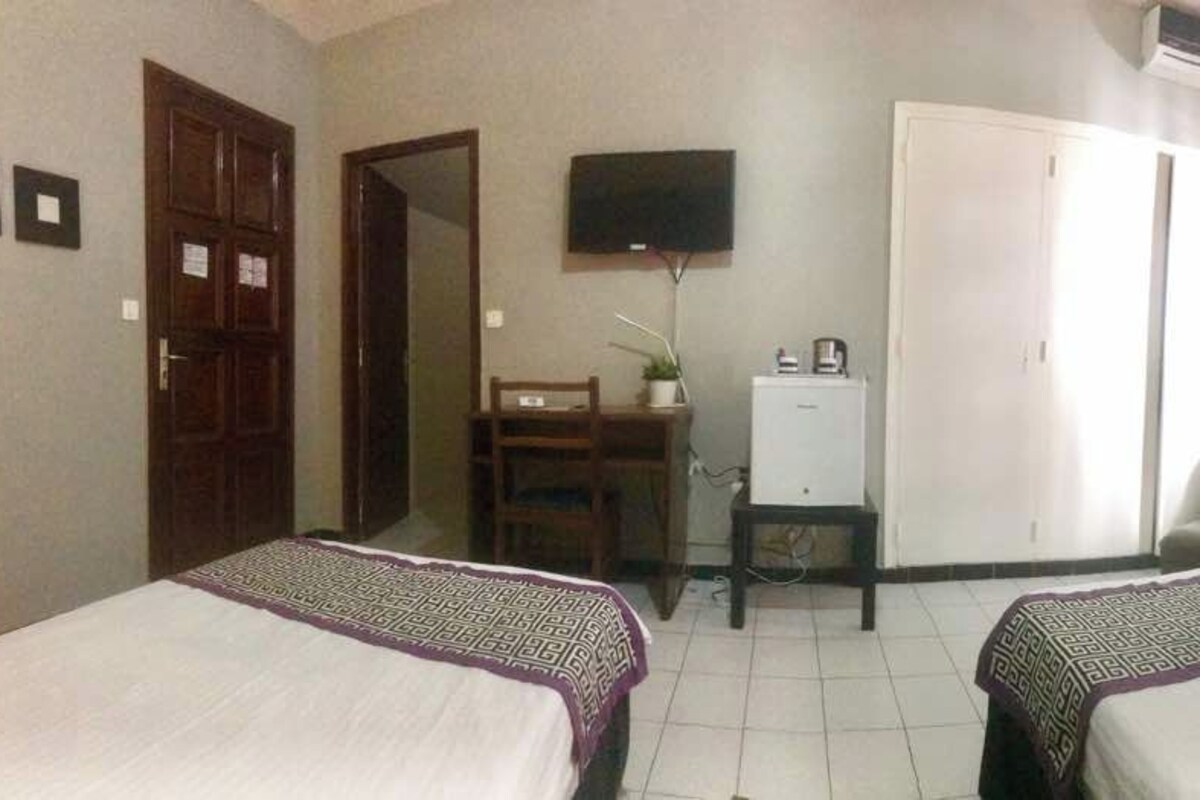 Ein Hotelzimmer mit zwei Betten, einem Schreibtisch mit Computer, einem Fernseher, einem Kleiderschrank und einem Sessel.