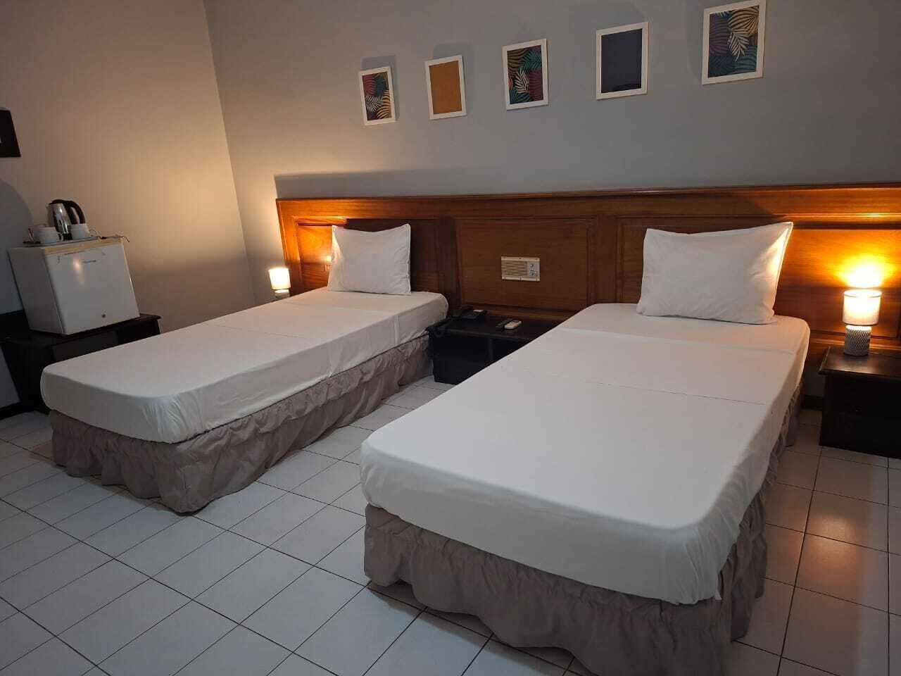A hotel room with two single beds, a wooden headboard, bedside lamps, a small table with a kettle, and a refrigerator.
