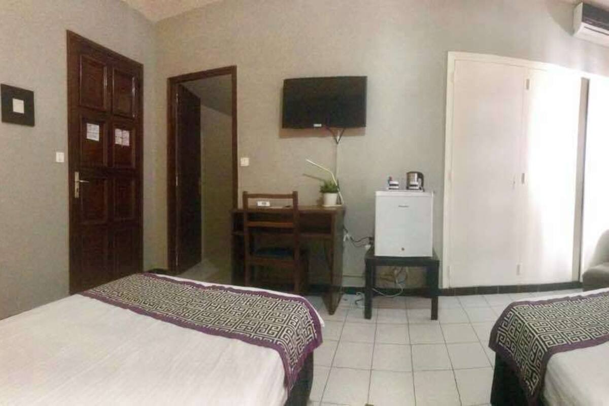 Une chambre d’hôtel avec deux lits, un bureau avec un ordinateur, une télévision, une armoire et une chaise.