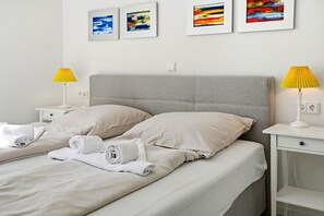 A neatly made bed with pillows and rolled towels, two bedside tables with lamps, and four framed abstract paintings on the wall. - Family & friends - Holiday apartment family & friends: balcony, elevator & great (Norden)