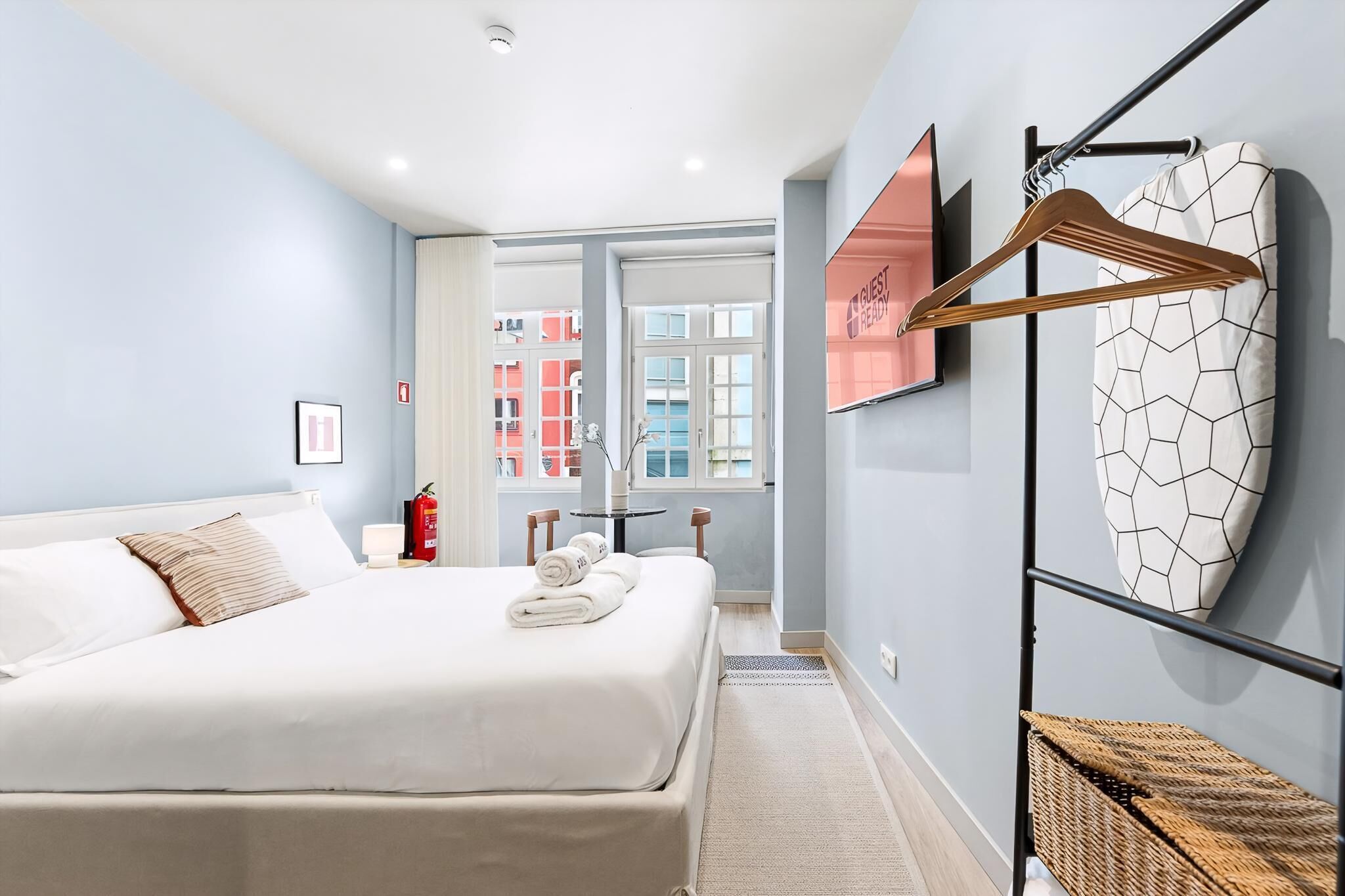 Hotelkamer met een bed, een wandtelevisie, een brandblusser, een raam en uitzicht op gebouwen.