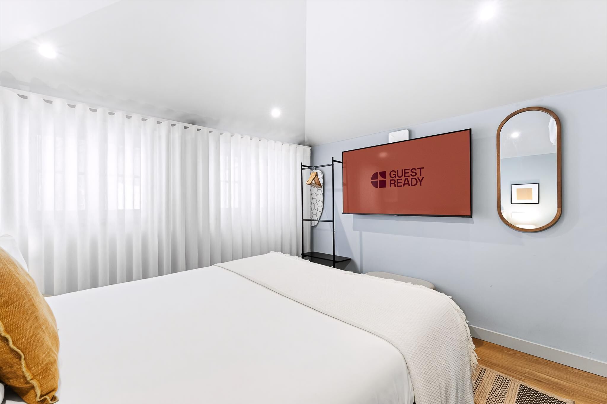 Ein modernes Hotelzimmer mit einem Bett, einem an der Wand befestigten Fernseher, einem Spiegel und einem Fenster mit weißen Vorhängen.