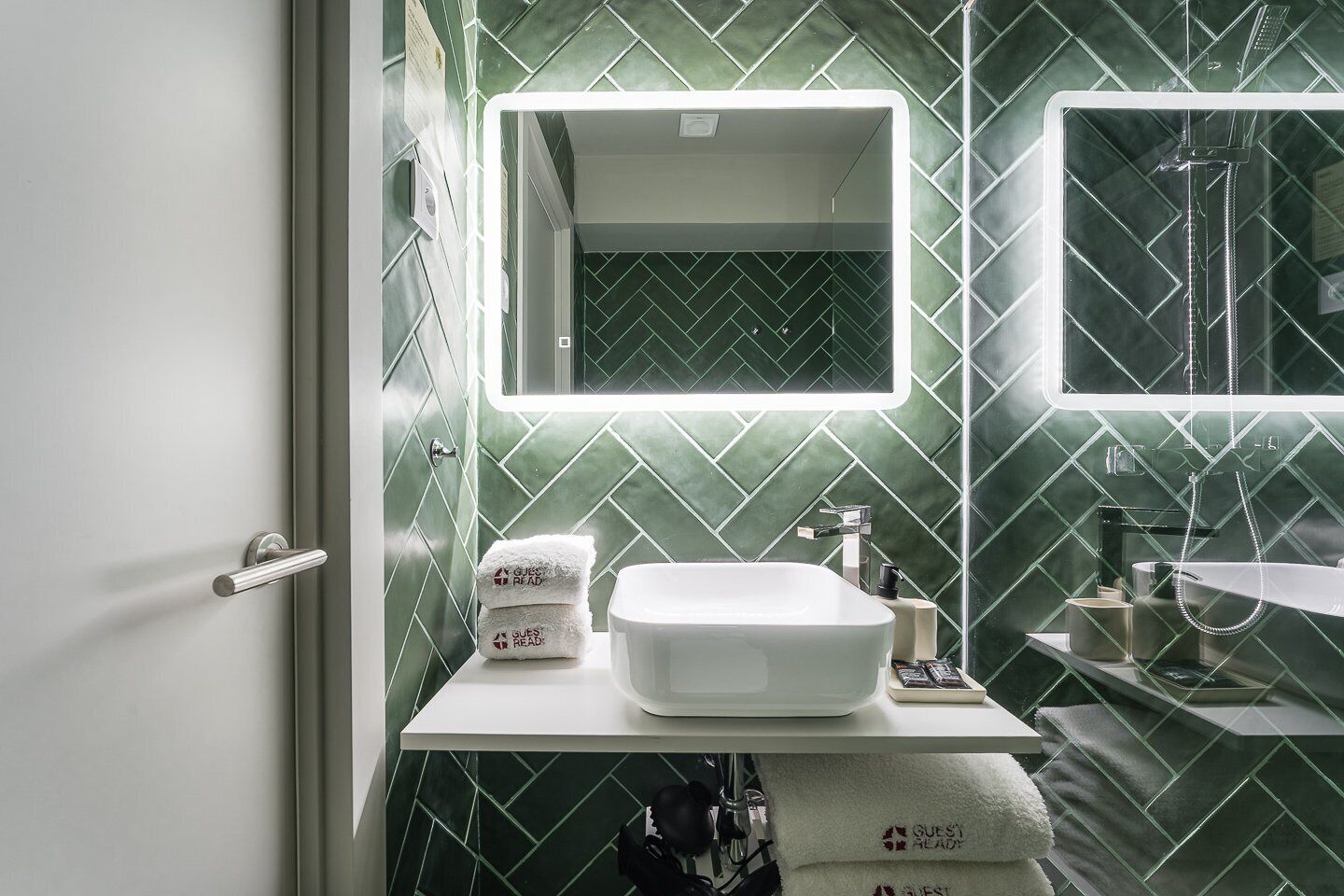 A bathroom with a white sink, a mirror, and green patterned tiles. There are towels on the counter and a shower area visible in the mirror.