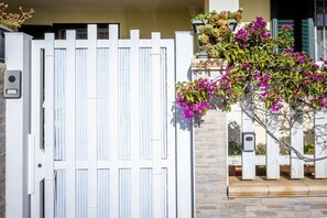 A white gate with vertical slats, a stone wall, and a flowering plant with pink flowers. - GuestHost - Relax & Fun Home between sea & city (Sinnai)