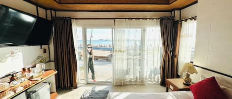 A hotel room with a bed, a desk, a TV, a chandelier, and a view of the beach.