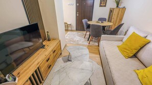A modern living room with a TV, wooden furniture, a marble coffee table, a sofa with yellow pillows, and a dining area with chairs and a table. - Urban Zen  Skopje Modern 1BR in Skopje Center ,Quiet & Fully Equipped (Skopje)
