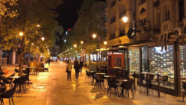 A nighttime street with outdoor seating, trees, and illuminated storefronts. - Urban Zen  Skopje Modern 1BR in Skopje Center ,Quiet & Fully Equipped (Skopje)
