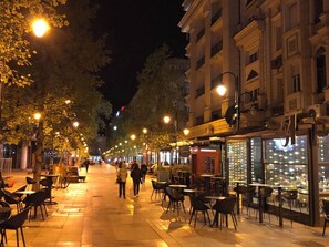 Une rue animée la nuit, avec des terrasses, des arbres et des devantures de magasins illuminées.