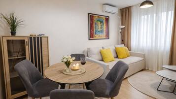 A modern living room with a round wooden table, two grey armchairs, a sofa with yellow cushions, a painting on the wall, and an air conditioning unit.