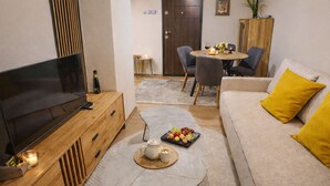 A living room with a TV, wooden cabinet, round coffee table with fruits, and a sofa with yellow pillows. - Urban Zen  Skopje Modern 1BR in Skopje Center ,Quiet & Fully Equipped (Skopje)
