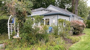 Ein zweistöckiges Haus mit grauer Fassade, umgeben von einem gepflegten Garten mit verschiedenen Sträuchern und einem kleinen Brunnen.