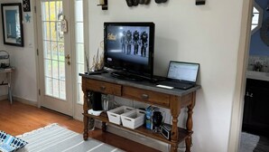 Ein Wohnzimmer mit einem Holztisch, einem Fernseher, der ein Spiel zeigt, und einem Schild mit der Aufschrift 'BEACH HOUSE' an der Wand.