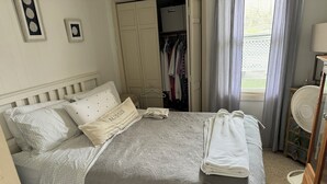 Ein ordentlich bezogenes Bett mit einer grauen Decke und Kissen, ein Nachttisch mit Lampe, ein Kleiderschrank und ein Ventilator in einem Zimmer mit Fenster und Vorhängen.