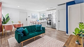 Eine moderne Wohnung mit einem türkisfarbenen Sofa, rosafarbenen Sesseln, einem Holzesstisch und einer Küche mit weißen Schränken und Edelstahlgeräten.