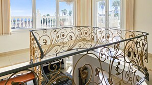 Un vaste balcon doté de balustrades en métal ouvragé et d’un lustre, offrant une vue sur une zone côtière bordée de palmiers.