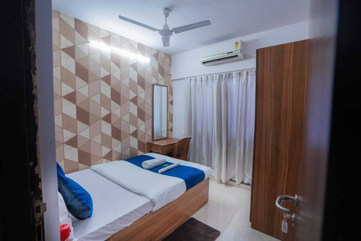 A bedroom with a bed, a ceiling fan, an air conditioning unit, a wooden wardrobe, and a mirror on the wall.