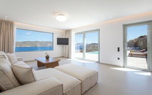 A modern living room with a large sofa, a coffee table, and a TV. It features large windows with a view of the sea and hills. - Villa Sozon. Spacious villa with sea view 150 m from the beach. (Mykonos)