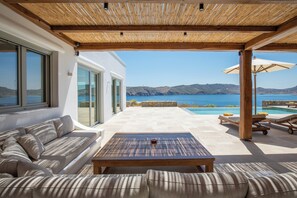 A modern outdoor patio with a wooden pergola, a sofa, a table, and a pool area with lounge chairs and an umbrella. - Villa Sozon. Spacious villa with sea view 150 m from the beach. (Mykonos)