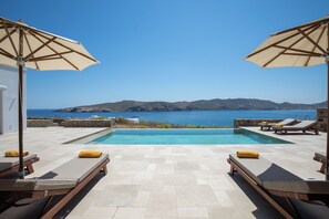 A pool with lounge chairs and umbrellas, a clear view of the sea, and a mountainous landscape in the background. - Villa Sozon. Spacious villa with sea view 150 m from the beach. (Mykonos)