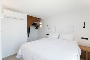 A bedroom with a bed, white bedding, a wooden bench, a wall-mounted light, and a ceiling fan. - Villa Sozon. Spacious villa with sea view 150 m from the beach. (Mykonos)